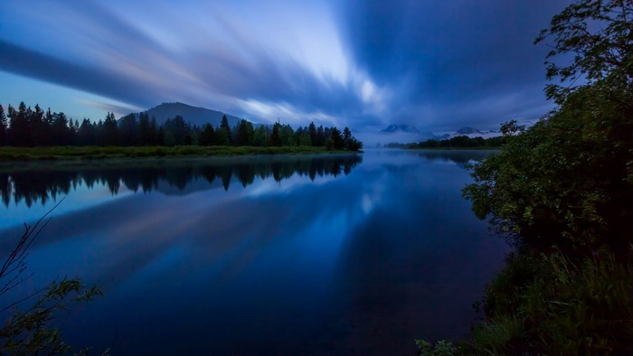 Long exposure of a river reflecting trees and mountains at dusk