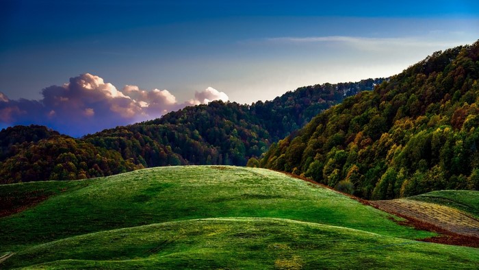 Rolling green hills and forested mountains under a blue sky with clouds