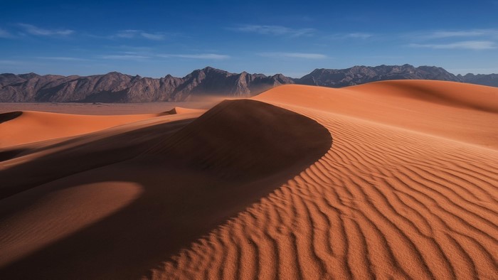 Sand dunes and desert mountains under a blue sky