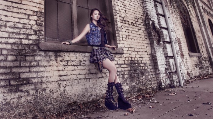 Girl in denim vest and plaid skirt leaning on brick wall