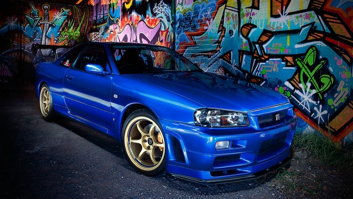 Blue Nissan Skyline R34 GT-R parked in front of graffiti wall