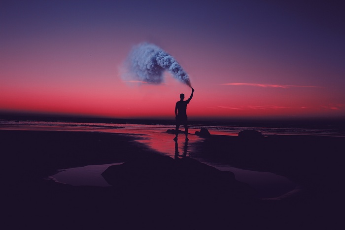 Silhouette of a person holding a smoke flare on a beach at sunset