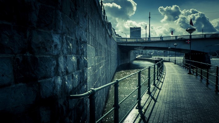 Stone wall and walkway along a river under a bridge