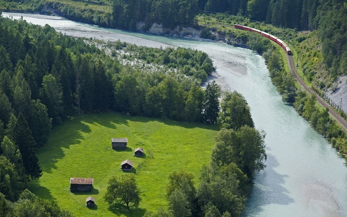 Red train travels along a river in a mountainous landscape