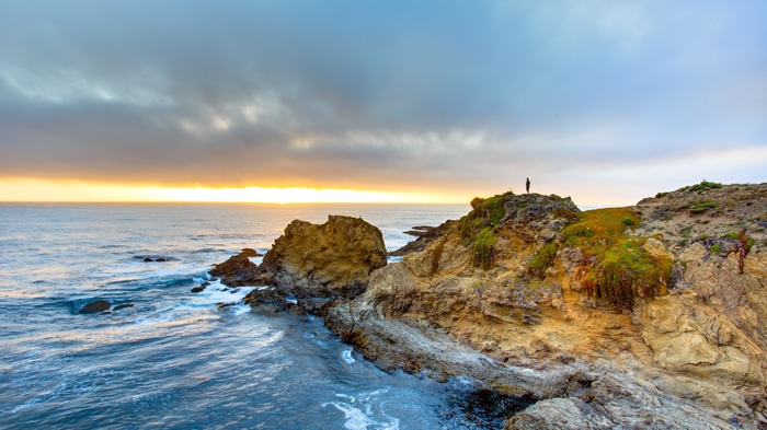 Person standing on a rocky cliff overlooking the ocean at sunset