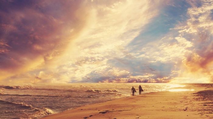 Two surfers walk along a beach at sunset