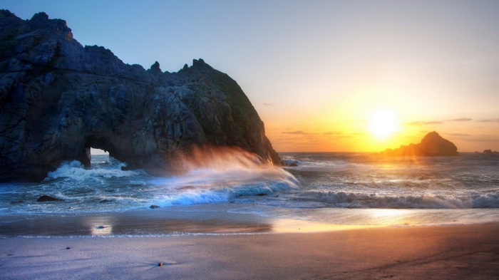 Waves crashing against a rock arch at sunset