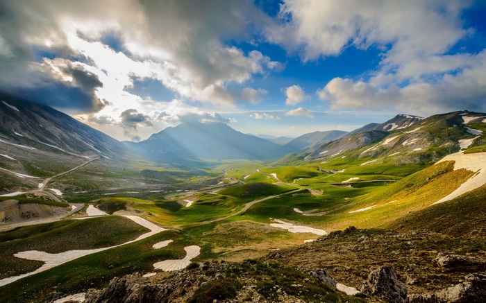 Sunlight streams through clouds over a green mountain valley with snow patches