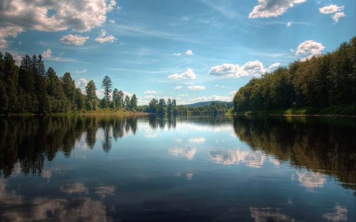 A wide river reflects trees and clouds under a blue sky.
