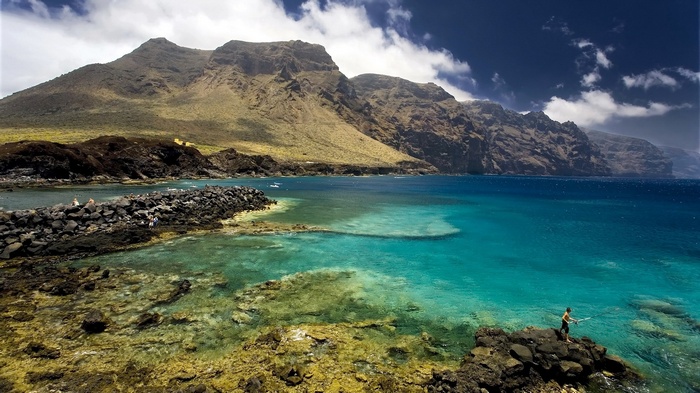 Man fishing on rocky coast with mountains and ocean