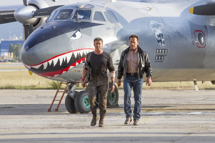 Sylvester Stallone and Arnold Schwarzenegger walk past an AN-26 airplane