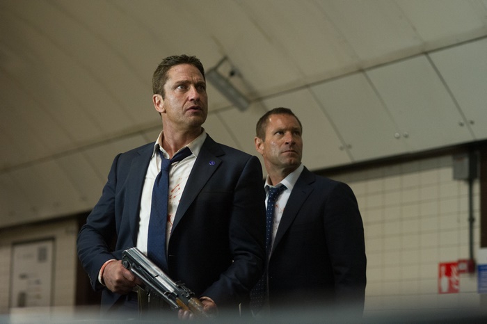 Gerard Butler in a dark suit holding a rifle