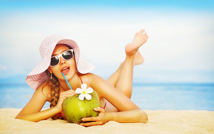 Woman in hat and sunglasses drinks coconut on beach