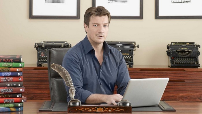 Nathan Fillion at a desk with typewriters and books