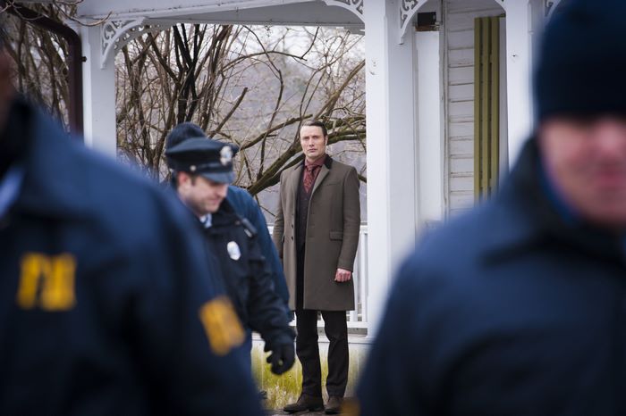 Mads Mikkelsen in a coat standing outside a house