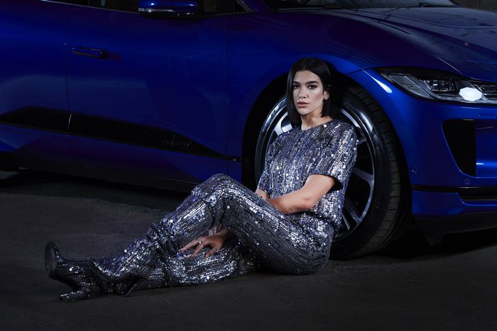 Dua Lipa sitting by a blue Jaguar car