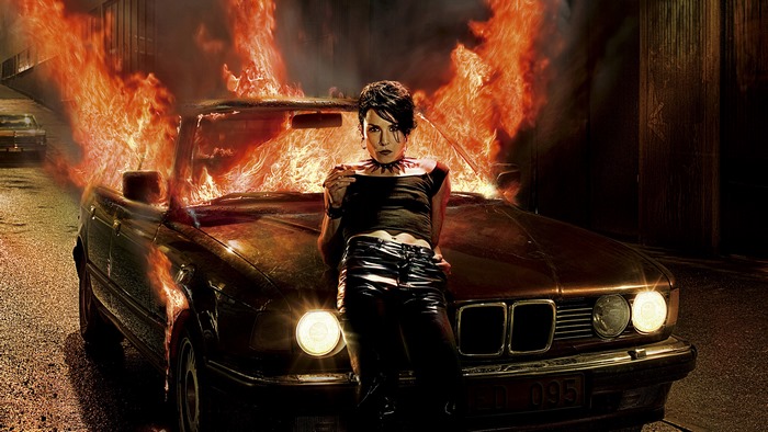 Noomi Rapace in front of a burning car