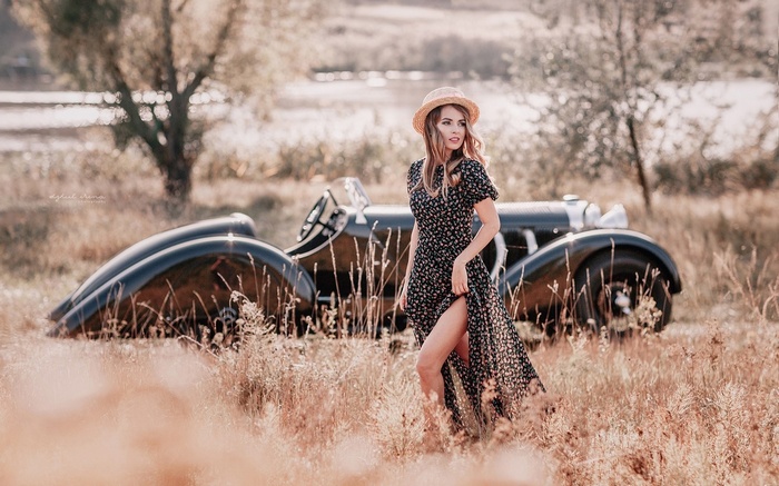 Woman in floral dress and straw hat posing by vintage car