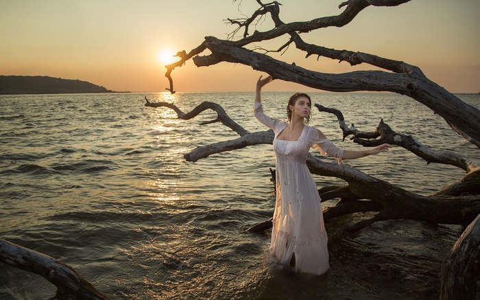 Woman in white dress standing in water near driftwood at sunset