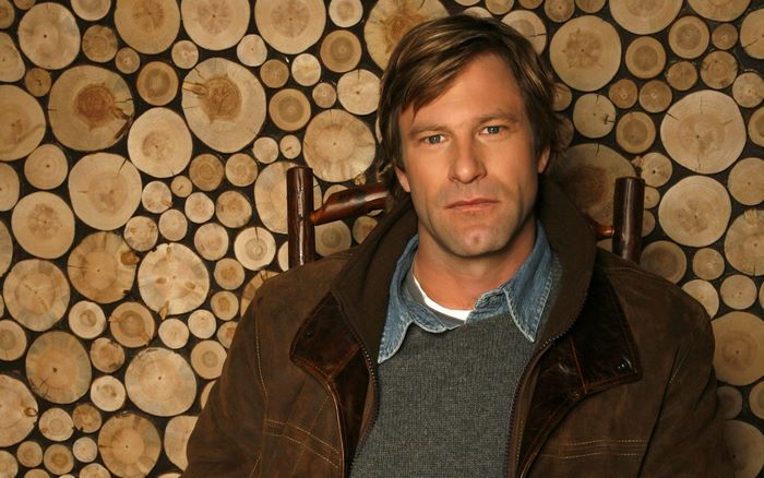 Aaron Eckhart sitting in front of a wood-slice wall