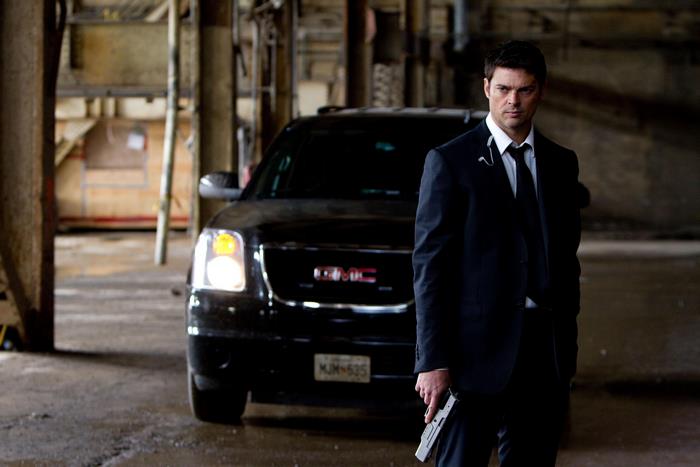 Karl Urban holding a gun in front of a GMC SUV