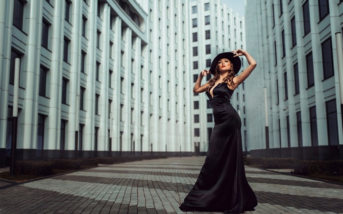 Woman in black dress and hat posing near modern buildings