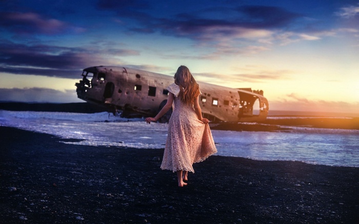 Woman in dress walks towards crashed airplane on beach at sunset