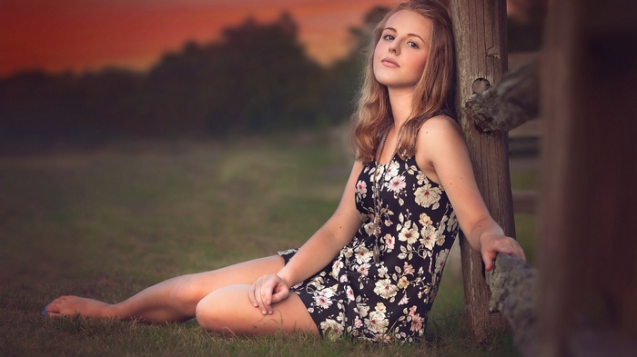 Young woman in floral dress leaning on wooden fence at sunset