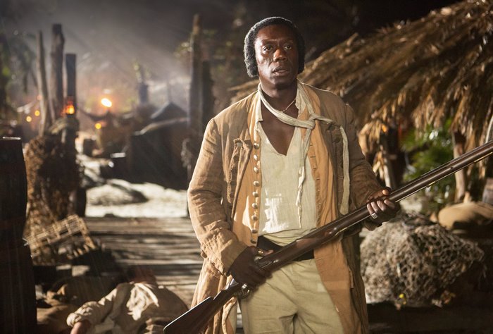 Hakeem Kae-Kazim holding a rifle in a village