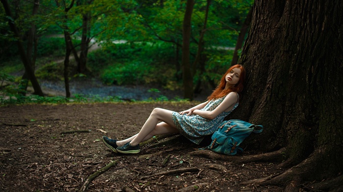 Red-haired girl sitting by a tree in a forest