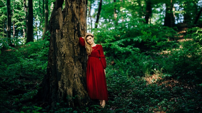 Woman in red dress leaning against a tree in a forest