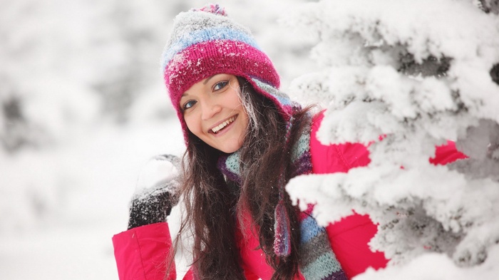 Young woman in winter clothing peeking from behind a snow-covered tree