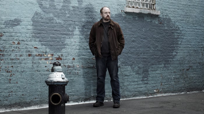 Louis C.K. standing by a brick wall and fire hydrant