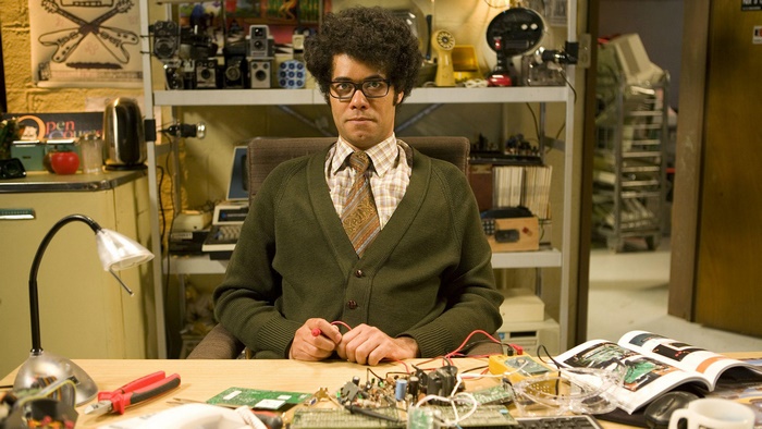 Richard Ayoade sitting at a desk with electronics