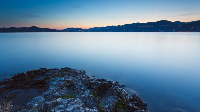 Rocky shore at dusk overlooking a calm lake and distant mountains with city lights