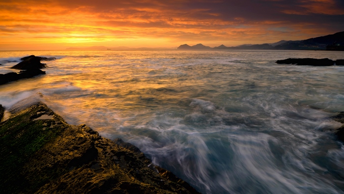 Sunset over the ocean with waves crashing on rocks