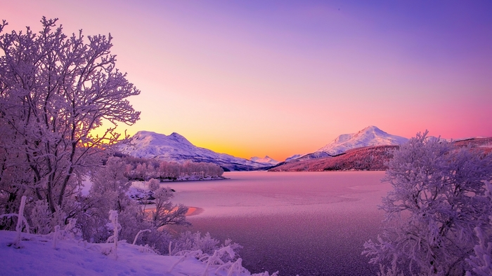 Winter sunset over a frozen lake with snow-covered mountains and trees