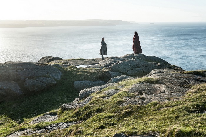 Two figures stand on a rocky coastline overlooking the sea