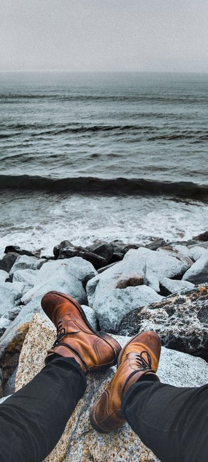 Person wearing boots and black jeans sitting on rocks by the ocean