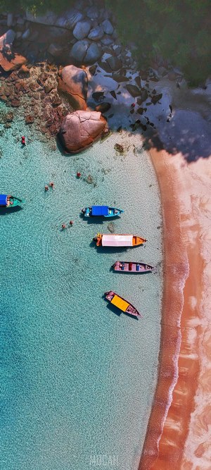 Aerial view of boats and people swimming in clear turquoise water near a sandy beach with large rocks.