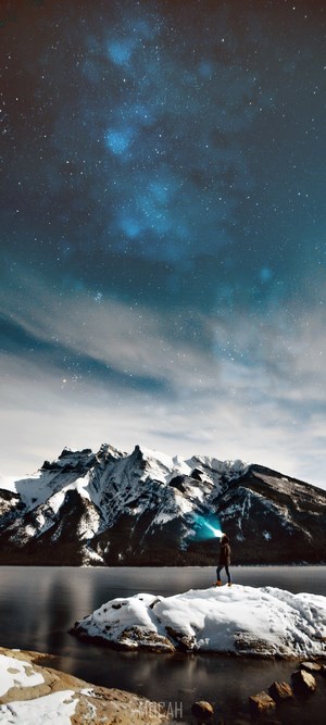 Person with flashlight on snowy rock by lake and mountains under starry sky
