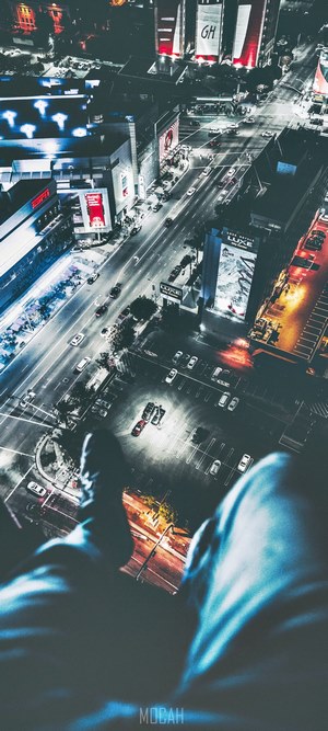 Person dangling legs off building roof overlooking city at night