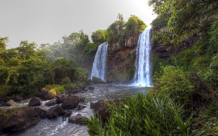 Two waterfalls cascade down rocky cliffs into a river surrounded by dense green foliage.