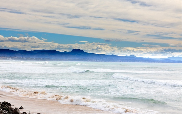Ocean waves crash on a sandy beach with mountains in the distance.
