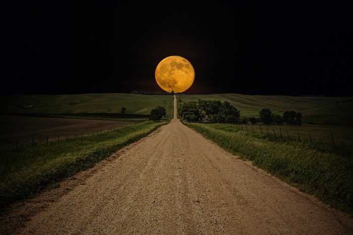 Dirt road leading to a full moon over rolling hills
