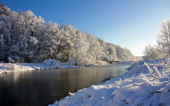 Snow-covered trees line a frozen river under a clear blue sky