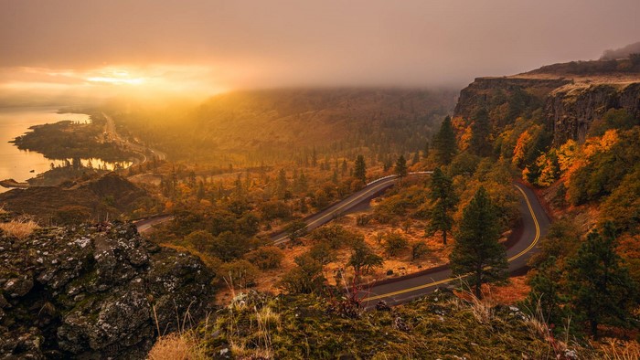 Winding road through autumn forest with river and sunset