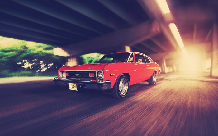 Red 1970s Chevrolet Nova driving under a bridge