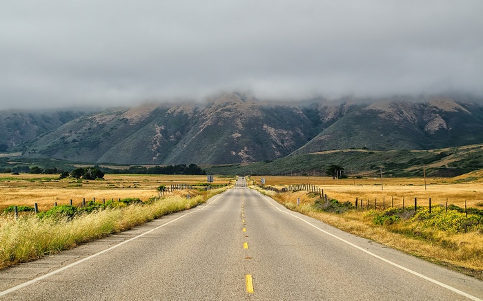 Empty highway through a valley towards mountains under clouds