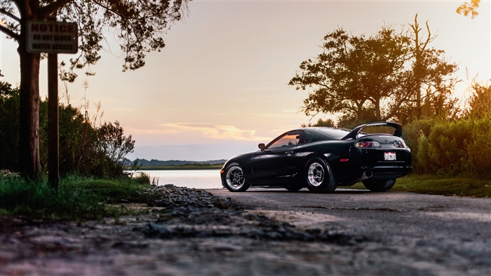 Black Toyota Supra parked by water at sunset